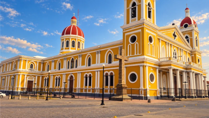 Yellow-and-white Granada Cathedral in Nicaragua with red-and-white domes and a stone cross in the plaza.