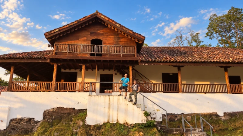 Two visitors on the steps of La Casona, a white-walled historic building with a wooden balcony and tiled roof beneath a blue sky.