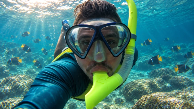 Snorkeler wearing a black mask and yellow snorkel above a rocky seabed, with another swimmer in the background.